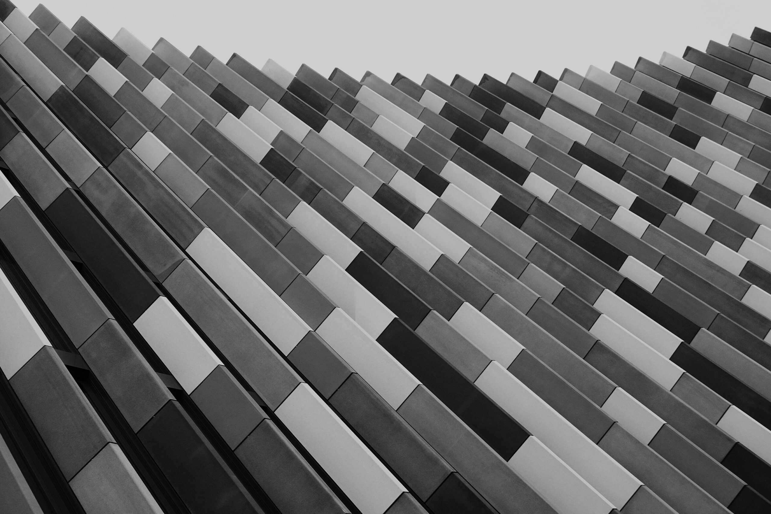 A monochrome image of a modern architectural facade composed of overlapping rectangular panels, creating a dynamic, wave-like pattern. The panels vary in shades of gray, adding depth and texture to the design.