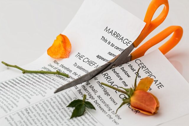 A marriage certificate is being cut in half with orange scissors. Two orange roses frame the scene, symbolizing separation. The image subtly hints at broader themes like equitable distribution, as assets and retirement funds are often divided in these life transitions; Division of Assets in New Jersey concept image