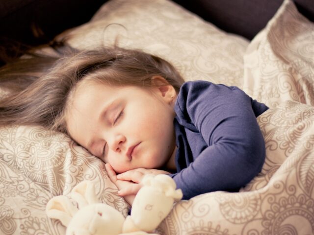 A young child with long hair sleeps peacefully on a bed, cuddling a stuffed animal while resting on a patterned pillow. The child wears a blue long-sleeved shirt. In this serene moment, the custodial parent finds solace amidst the challenges of divorce.
