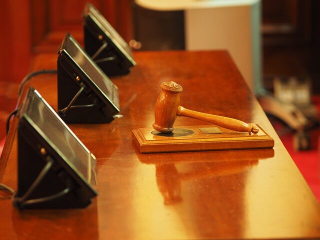 A wooden gavel rests on a matching sound block atop a polished table, hinting at the gravity of decisions made here. Nearby, three electronic devices with screens suggest serious discussions, possibly regarding a divorcing couple from different states in this formal judicial setting.