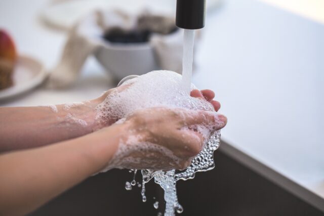 Hands covered in soap are being rinsed under running water at a sink, emphasizing hygiene's importance during coronavirus disease. In the background, a white bowl with dark contents and a piece of fruit are slightly blurred.