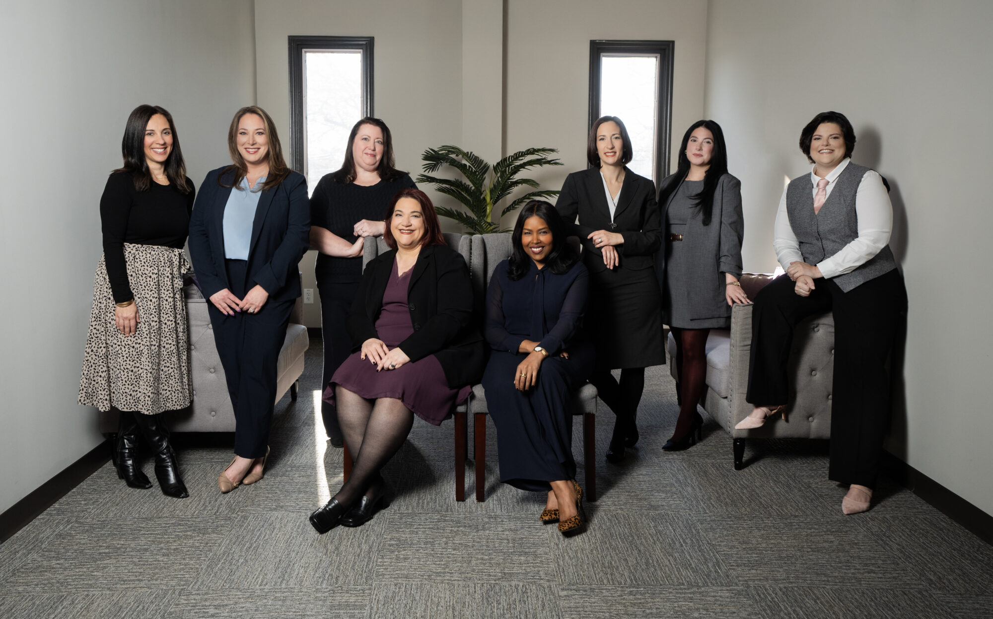 Eight women are posed in an office space with two armchairs. Some are standing, while others sit on or beside the chairs. They are dressed in professional attire. A potted plant is in the background between two windows.