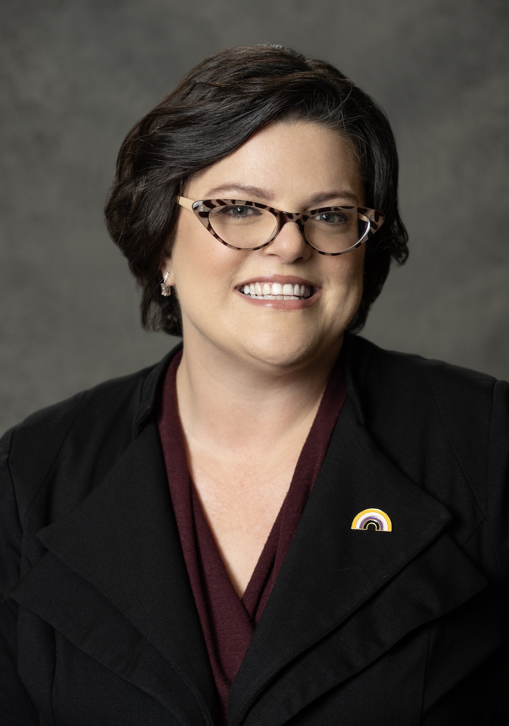 Jodi Argentino A person with short dark hair and glasses, wearing a black jacket and maroon top, smiles at the camera. They have a pin with a rainbow design on their lapel. The background is a plain, dark gray.