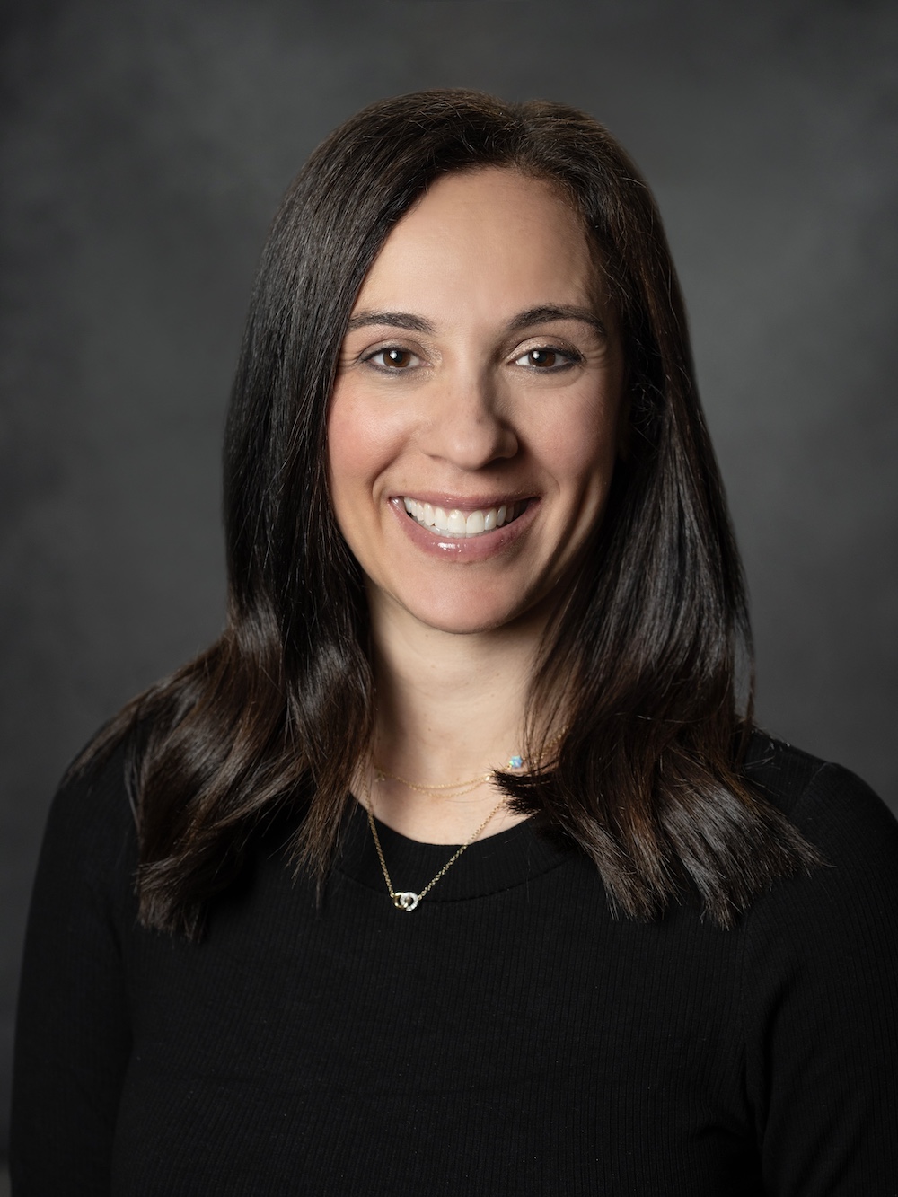 Allison Holzman A person with long dark hair, wearing a black top, smiles warmly at the camera against a dark background.