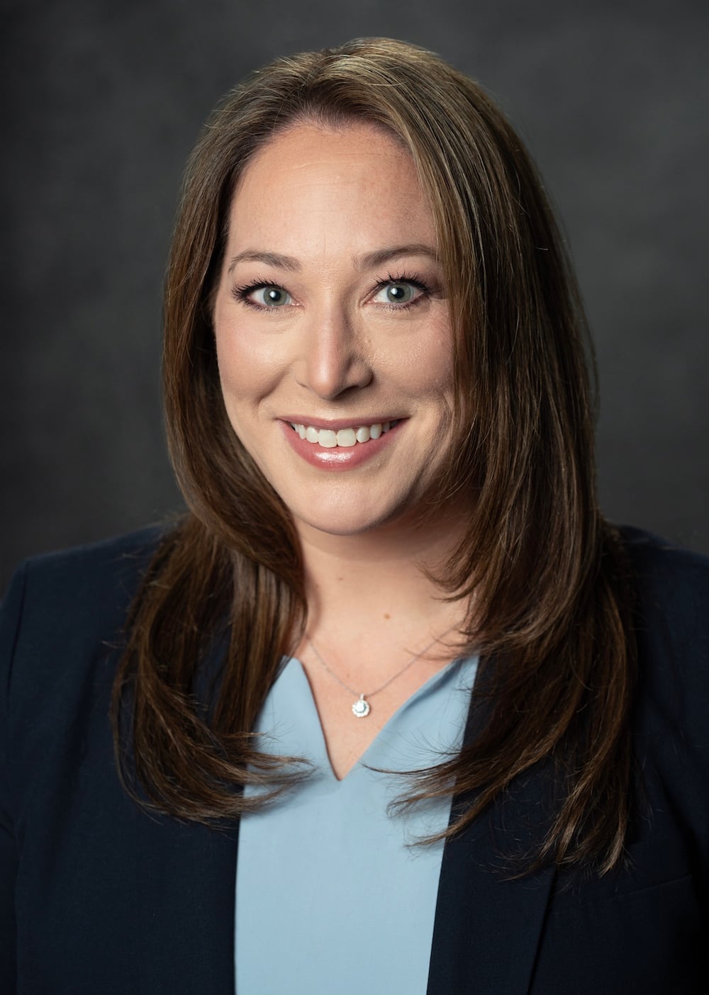 Portrait of Melissa E. Gluck, a smiling individual with long brown hair, donning a dark blazer over a light blue top. The background is dim and blurred, drawing attention to Melissa's welcoming expression.