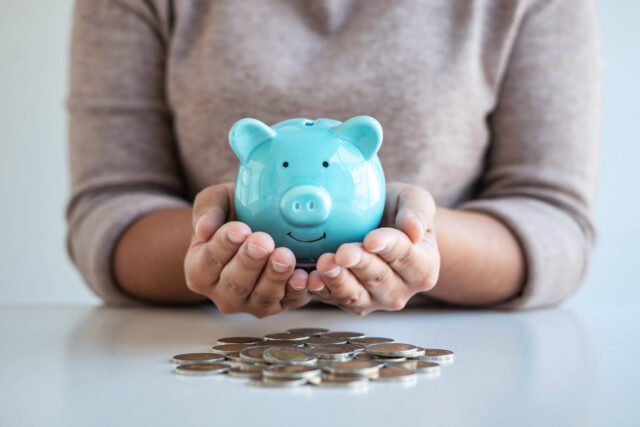 A person in a beige sweater holds a blue piggy bank with both hands, with a pile of coins spread out on a white table—symbolizing saving for retirement or managing spousal support in New Jersey.