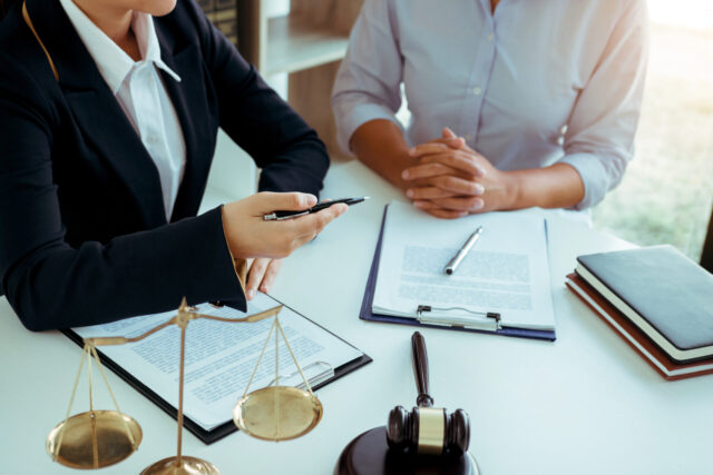 Two people sit at a desk with legal documents, a gavel, and scales of justice. One person gestures with a pen while the other listens, hands clasped, suggesting a legal consultation or meeting.