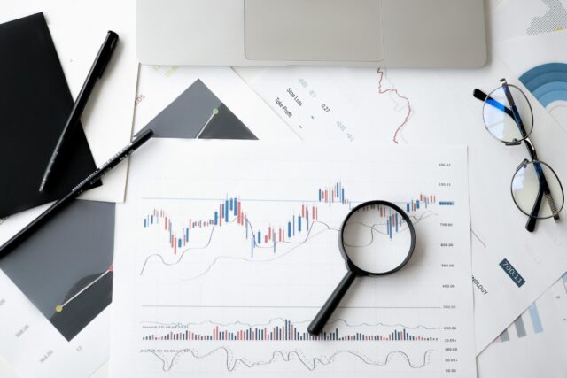 A desk with financial charts, a magnifying glass, glasses, pen, notebook, and a laptop suggests financial analysis or stock market research—ideal for tasks like financial disclosure during a divorce. The focus is on a chart with candlesticks and trend lines.