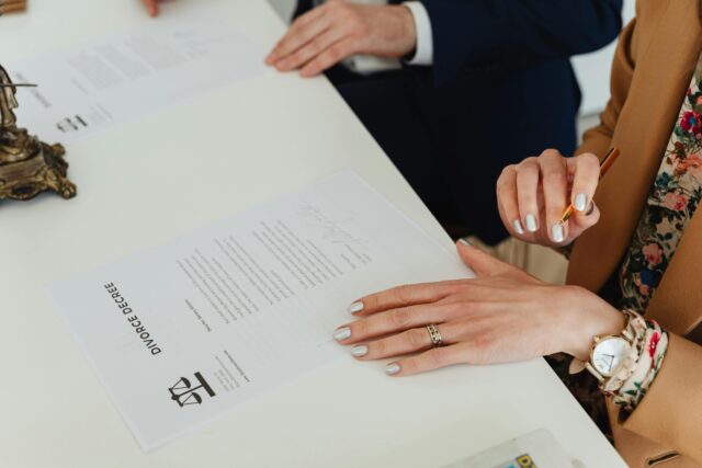 Two people sit at a table with divorce papers, highlighting how DIY divorces can lead to common mistakes. One person holds a pen, wearing a tan blazer and floral shirt, while the other’s hands rest near another document in the background.
