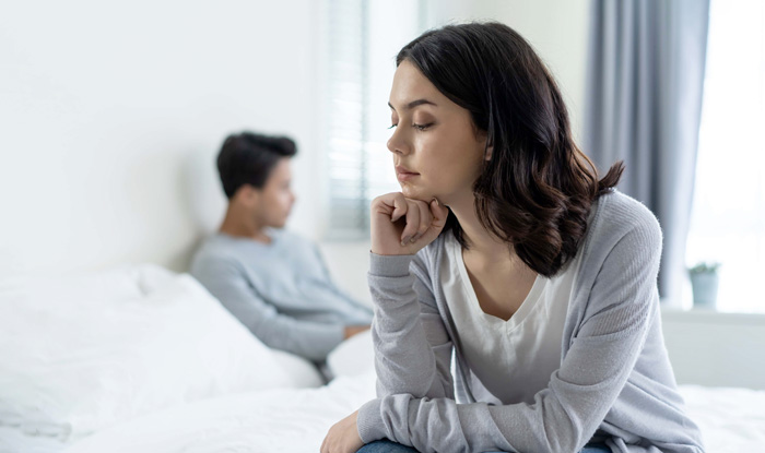 stressed woman sitting on edge of bed with husband in background