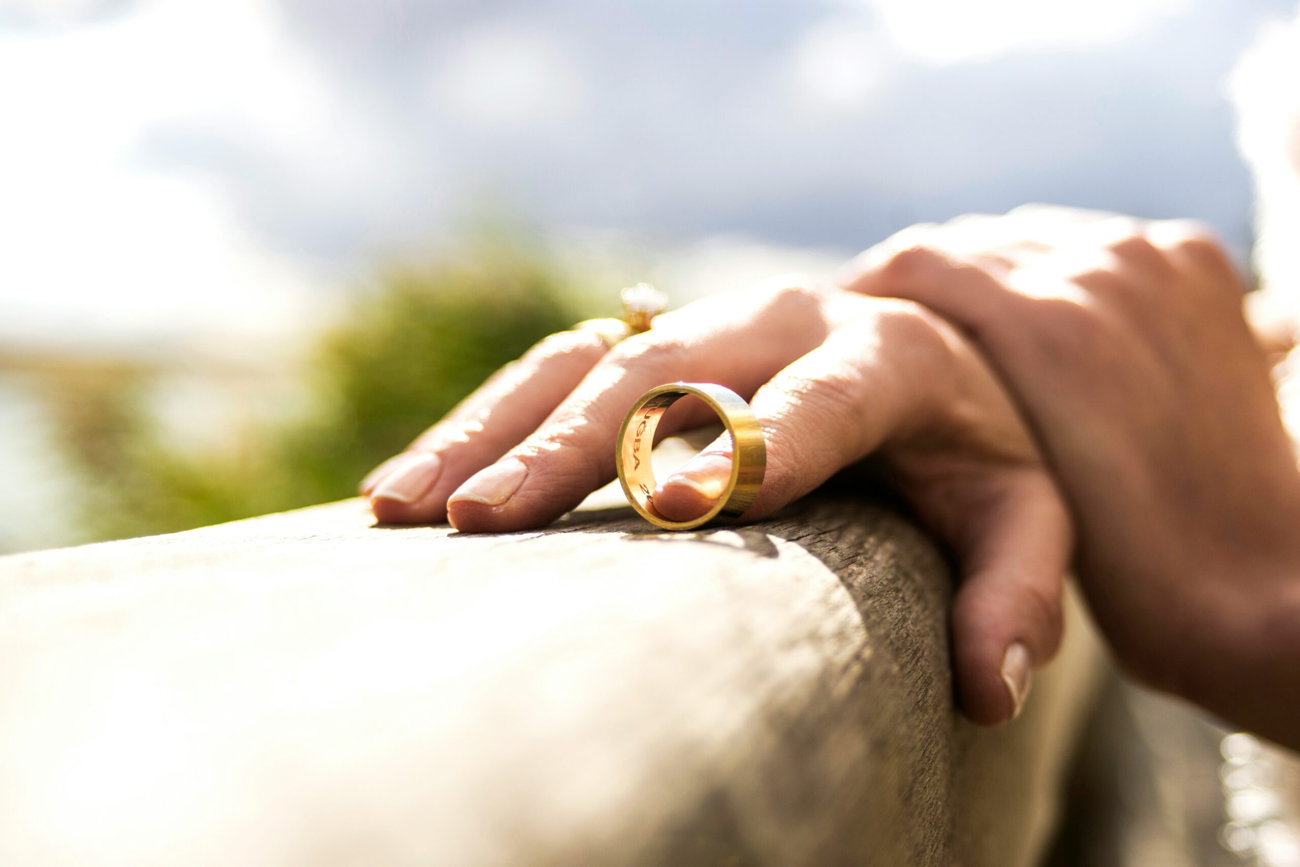 A hand with a gold ring, symbolizing memories and perhaps a separation agreement, rests on a wooden railing outdoors, sunlight reflecting off the ring against a blurred natural background.