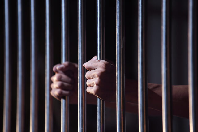 A close-up of two hands tightly gripping metal jail bars, with a dimly lit background, suggesting confinement or imprisonment—reflecting the struggles faced when a spouse is in prison during a New Jersey divorce.