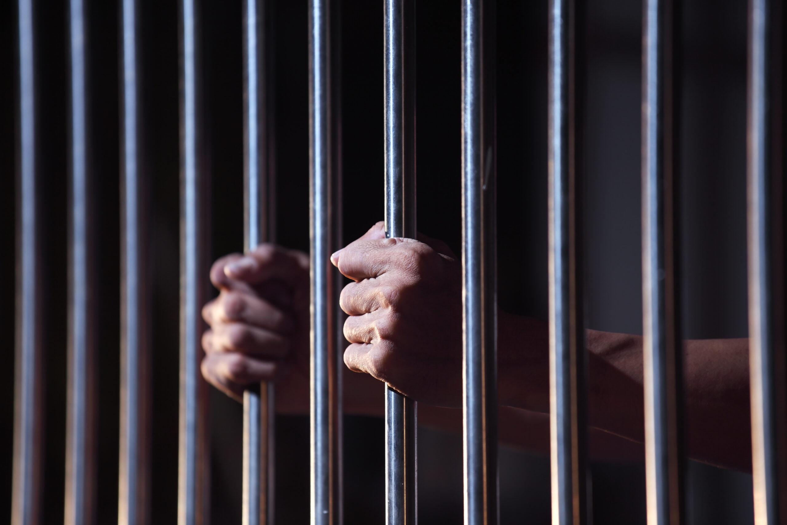 A close-up of two hands tightly gripping metal jail bars, with a dimly lit background, suggesting confinement or imprisonment—reflecting the struggles faced when a spouse is in prison during a New Jersey divorce.