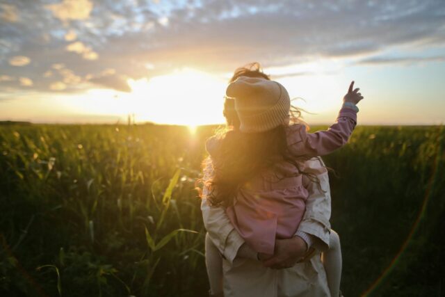 An adult carries a child through a grassy field at sunset, guided by the best interest of the child. The child wears a hat and points toward the bright sky, as sunlight creates a warm glow around them.