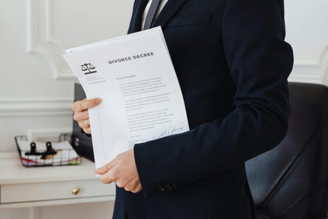 A person in a dark suit holds a divorce decree document in an office setting, with a desk and office supplies visible in the background.