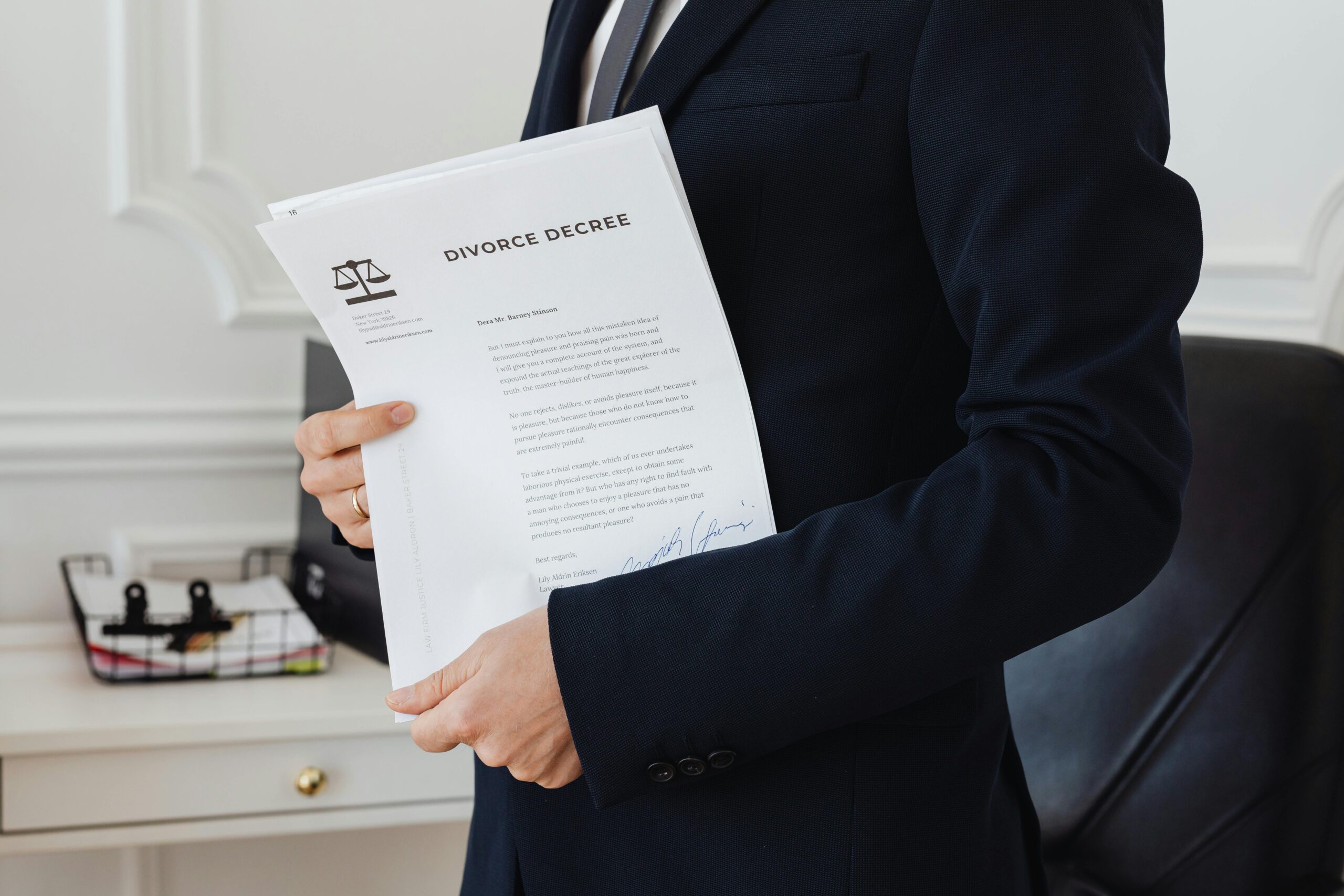 A person in a dark suit holds a divorce decree document in an office setting, with a desk and office supplies visible in the background.