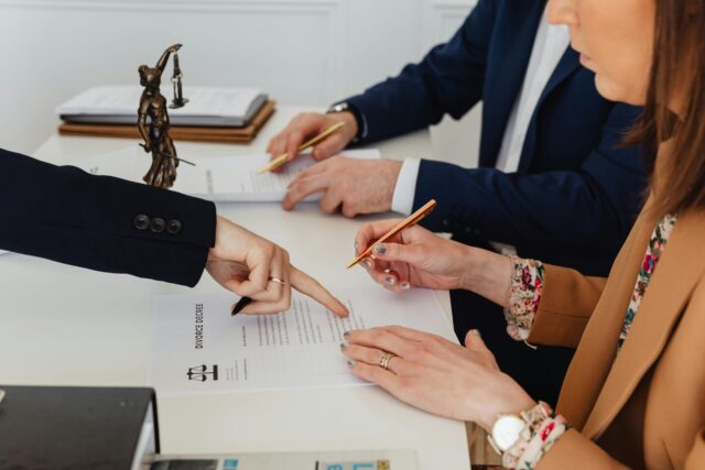 Three people sit at a table reviewing and signing collaborative divorce papers; one person points to a section on the document, while another holds a pen, and a small statue of Lady Justice is visible on the desk.