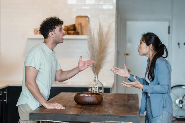A man and a woman stand on opposite sides of a wooden kitchen table, facing each other and arguing with expressive hand gestures. The atmosphere appears tense and emotional.